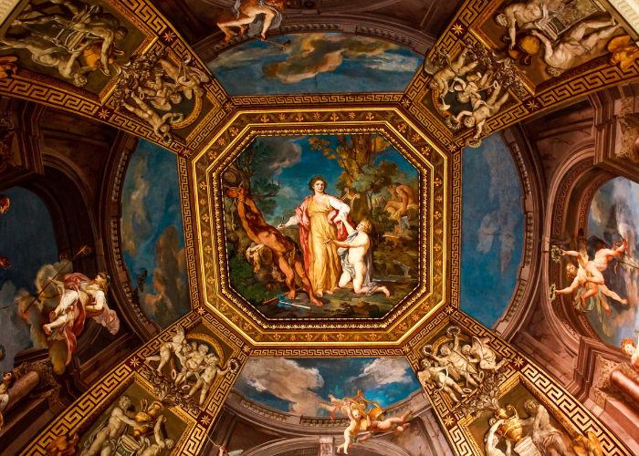 Photo of Renaissance Art painted on a ceiling. Art is of a woman standing between two men. One man is strapped to a tree, and the other is on his knees by the woman's feet.